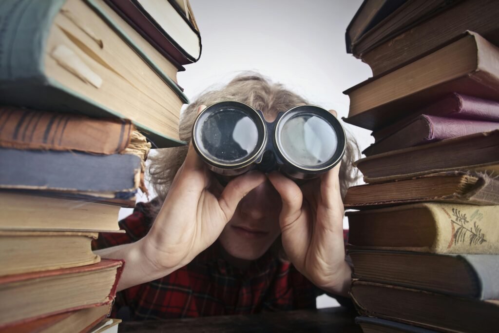 pexels-photo-3769697-3769697 A man observing through binoculars surrounded by stacks of vintage books indoors.